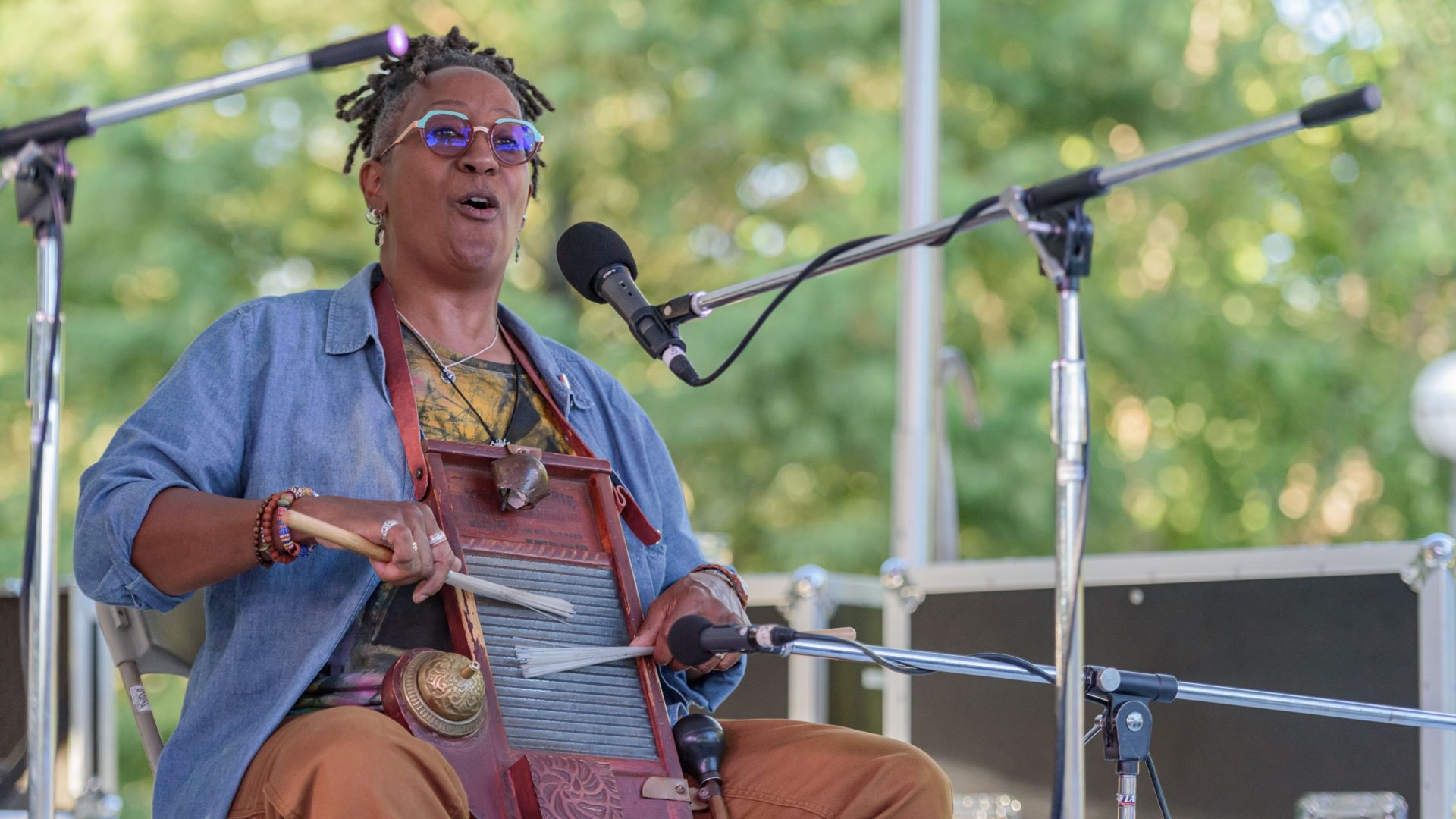 A seated musician performs on an outdoor stage, playing a washboard-style instrument mounted on the chest while singing into a microphone. The performer wears layered clothing and accessories, with multiple percussion elements attached to the instrument. Several microphone stands surround the performer, and leafy green trees are visible in the softly blurred background behind the stage.
