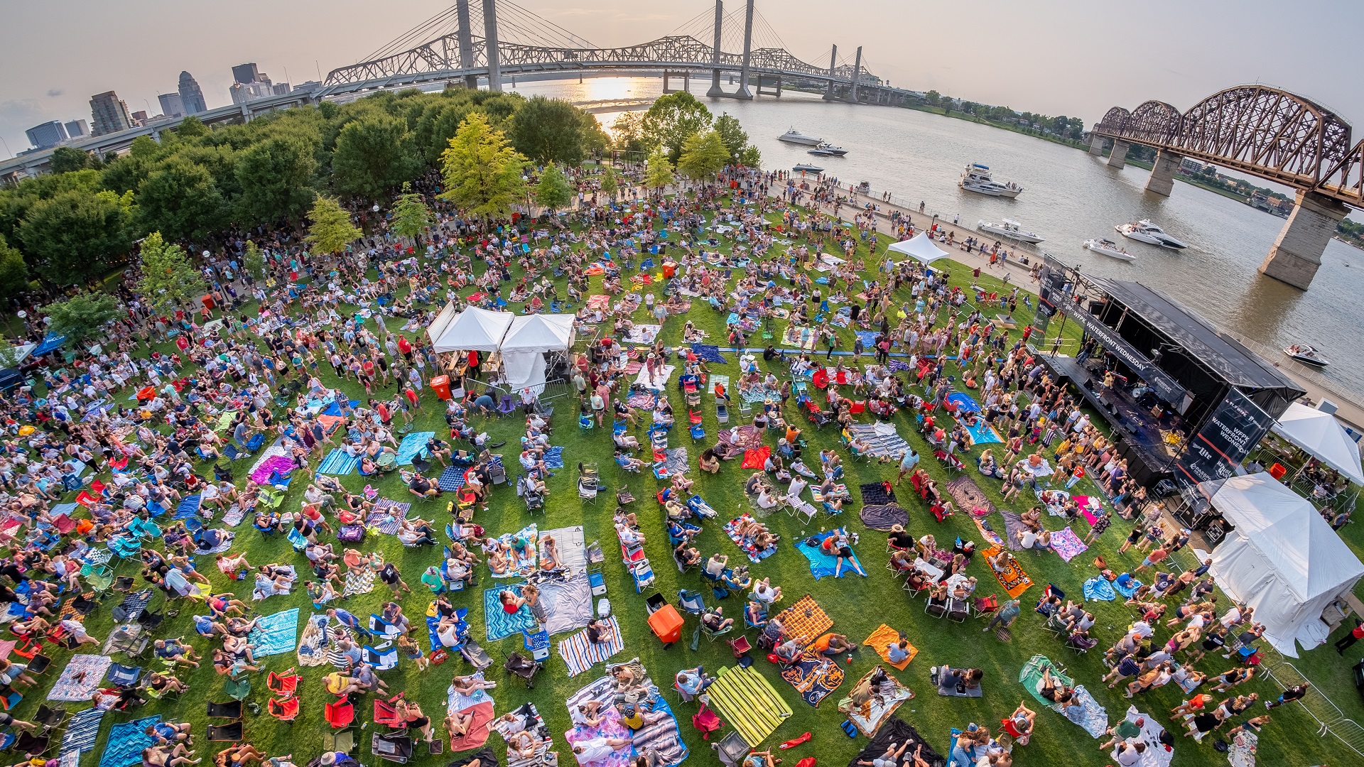 A large crowd sits on the Big Four Lawn watching live music with the Ohio River and Big Four Bridge as a backdrop.