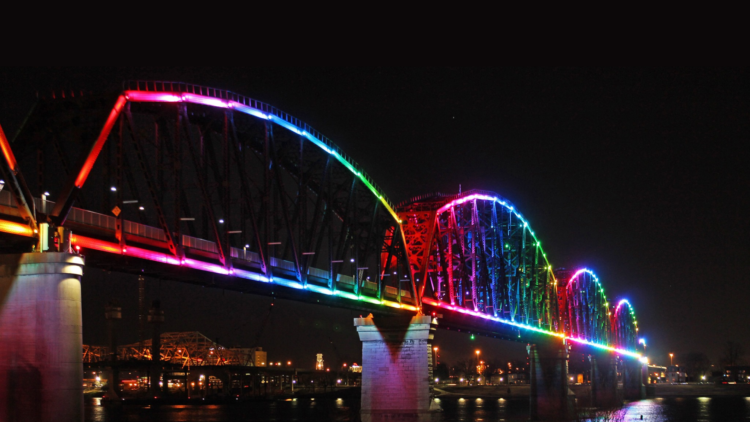 Big Four Bridge illuminated with rainbow LED lights at night, reflecting on river with city skyline.