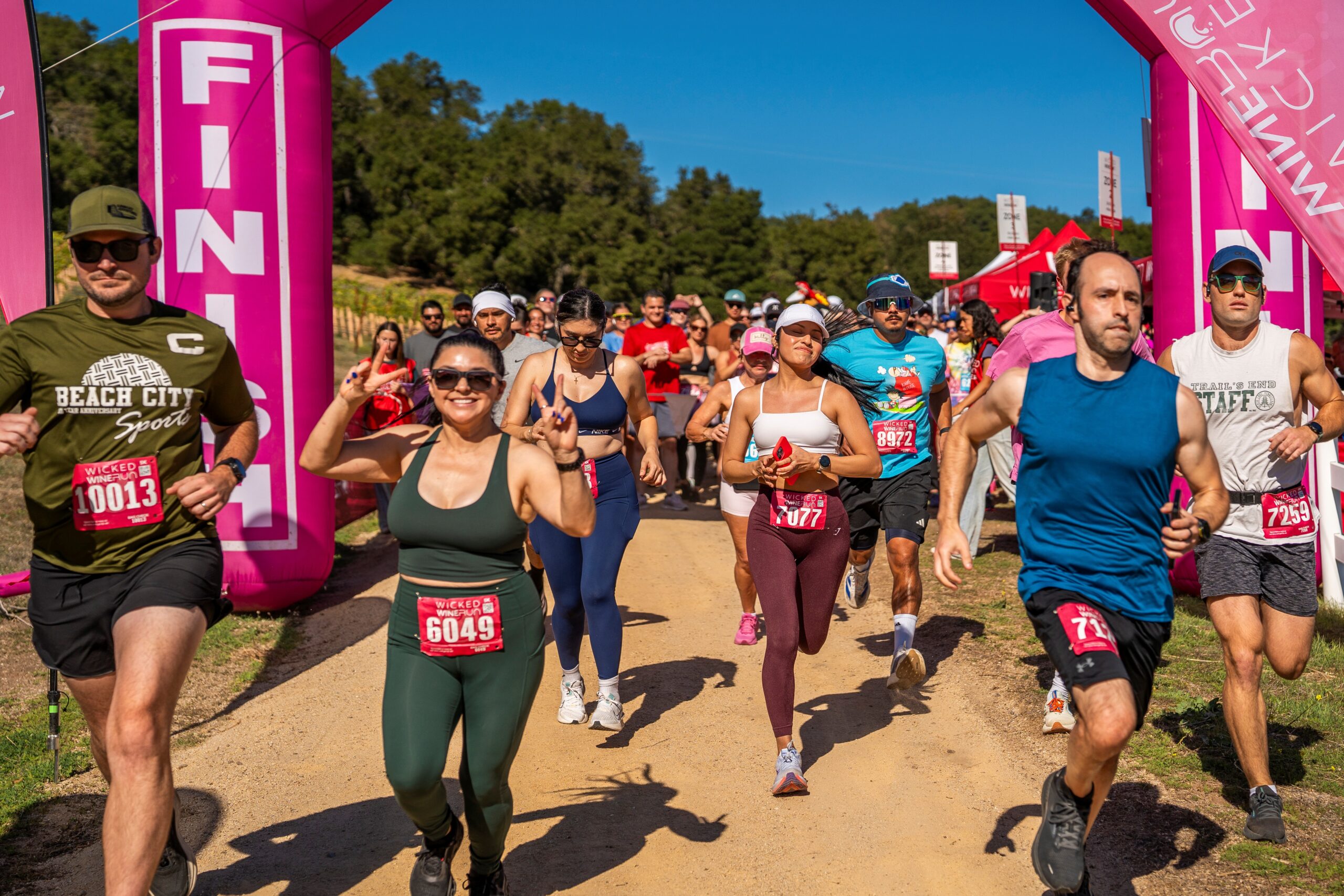 “Runners participating in an outdoor race as they leave the starting area, passing between bright pink event banners on a sunny day.”