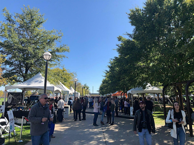 “People walking through an outdoor festival with vendor tents set up along a wide pathway on a sunny day, surrounded by green trees.”