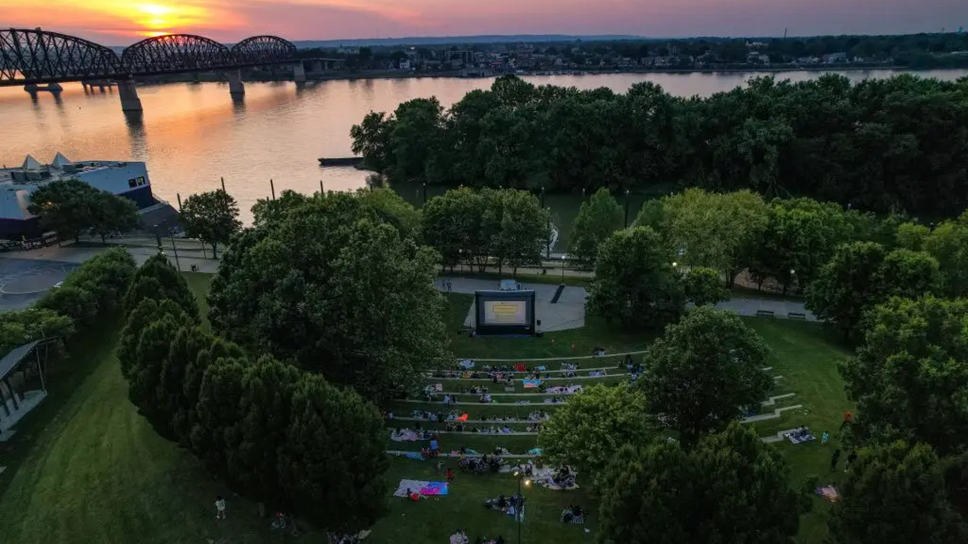 Aerial view of an outdoor movie screening in a riverside park at sunset, with attendees seated on a grassy amphitheater and a bridge spanning the river in the background.