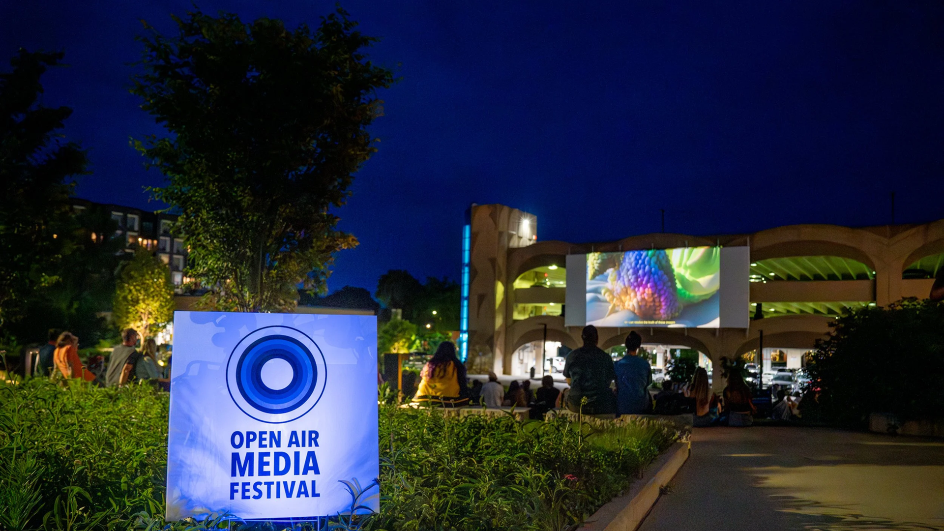 Nighttime outdoor scene of a crowd gathered in a grassy urban park, sitting on the lawn and low stone seating while facing a large building with a curved facade. A large digital projection screen on the building displays colorful visuals, illuminating the audience. Apartment buildings, trees, and walkway lighting surround the area, creating a lively community event atmosphere.