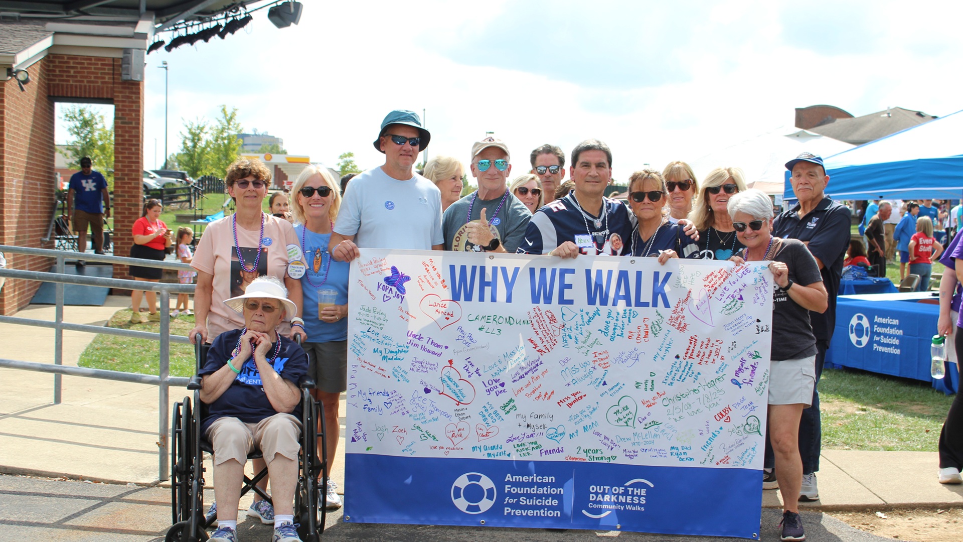 A group of participants, including one person using a wheelchair, hold a large “Why We Walk” banner covered in handwritten messages at an outdoor suicide‑prevention community walk.
