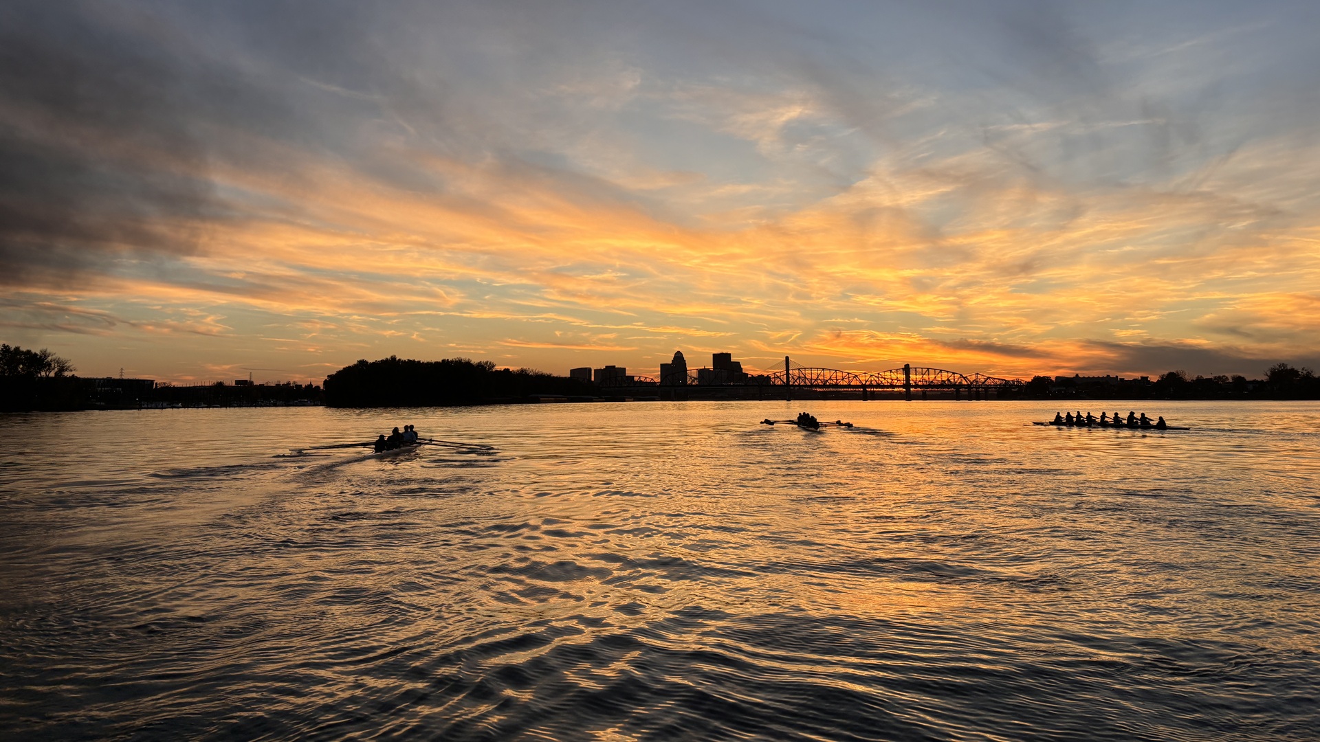 "Wide view of several rowing crews on a river during sunset. The sky is filled with orange, gold, and blue clouds, reflected across the water's surface. Silhouettes of rowers move across the river, while the Louisville skyline and a bridge appear on the horizon in the distance."